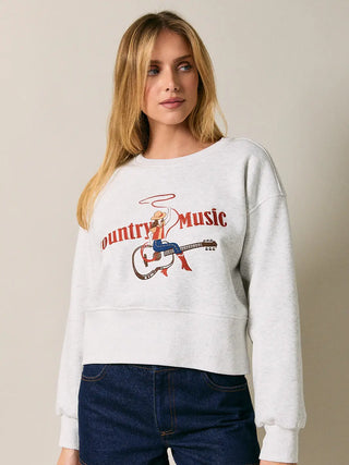 A woman wearing a light heather gray sweatshirt with a red and blue 'Country Music' graphic on the front, paired with dark blue jeans, against a solid beige background.
