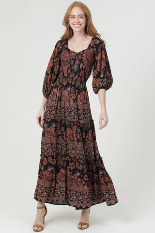 A woman modeling a long-sleeved maxi dress with a black and rust-red floral lace pattern, standing against a light gray studio background.
