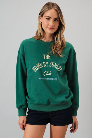 A woman wearing a green crewneck sweatshirt with cream-colored text that reads 'THE HOME BY SUNSET Club PRIVATE MEMBERS' on the front, paired with black shorts, against a solid gray studio background.