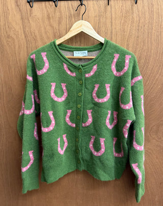 A green fuzzy knit cardigan sweater with pink glittery horseshoe patterns, hanging on a wooden hanger against a brown wood-paneled wall.