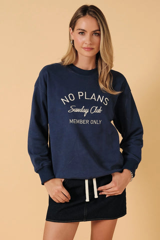 A woman modeling a navy blue crewneck sweatshirt with white text that reads 'NO PLANS Sunday Club MEMBER ONLY', paired with a dark denim skirt, against a solid beige background.