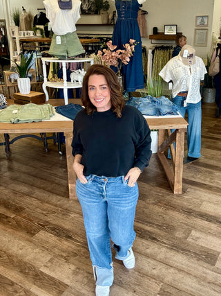 Woman standing in a clothing store wearing a black sweatshirt and cuffed blue jeans.