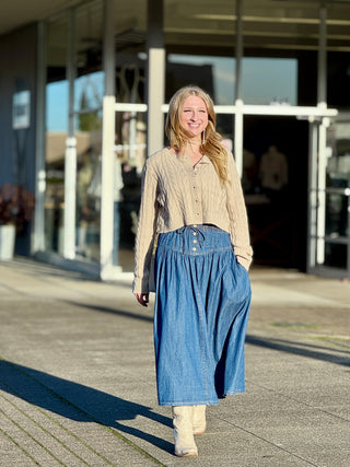 Woman wearing a beige cardigan and blue denim skirt standing on a sidewalk.